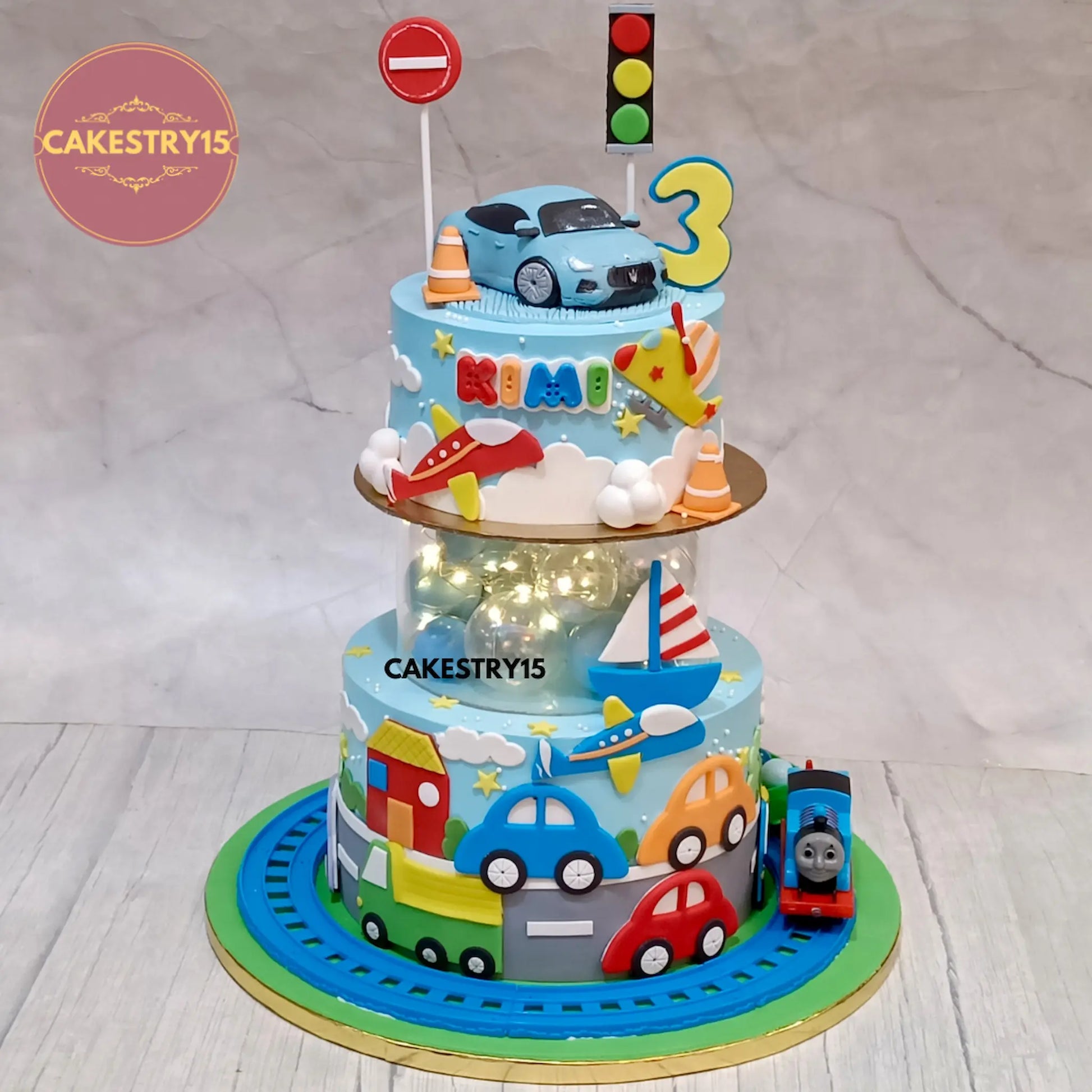 Two-tier birthday cake with spacer,toy cars, traffic lights, and a car on top, featuring the name Kimi and the number 3. Cake is branded with  CAKESTRY15