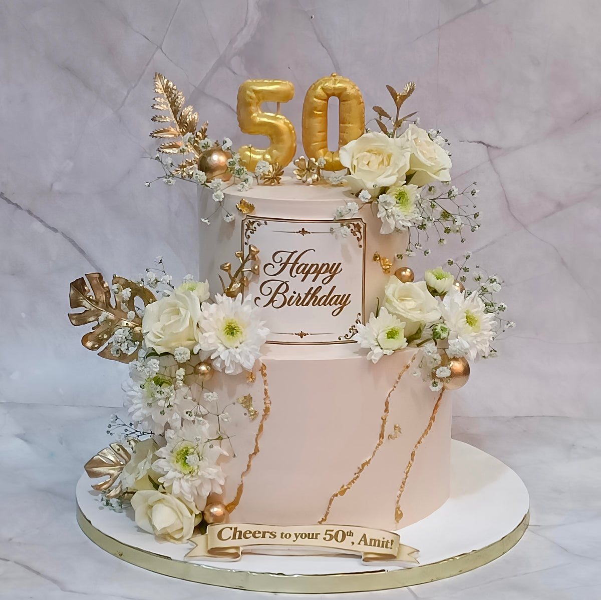 A two-tier birthday 50th birthday cake for men and women decorated with white and cream flowers, gold foil leaves, gold marble-like drizzle, and gold balloon numbers '50' on top. The front plaque reads 'Happy Birthday' and the base banner says 'Cheers to your 50th Amit'.