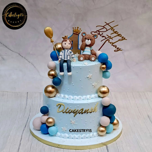 A 3kg, 2-tier chocolate teddy-themed birthday cake for Divyansh's first birthday, featuring a boy figurine with a crown, a teddy beside an age 1 tag, multi-colored faux balls, and a customized name tag by cakestry15,best bakery in noida