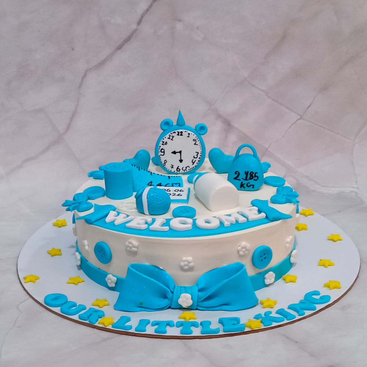 A custom baby shower cake decorated in blue and white with fondant details, featuring a clock, diaper, stethoscope, and a large bow on top, displayed on a white marble-patterned background.