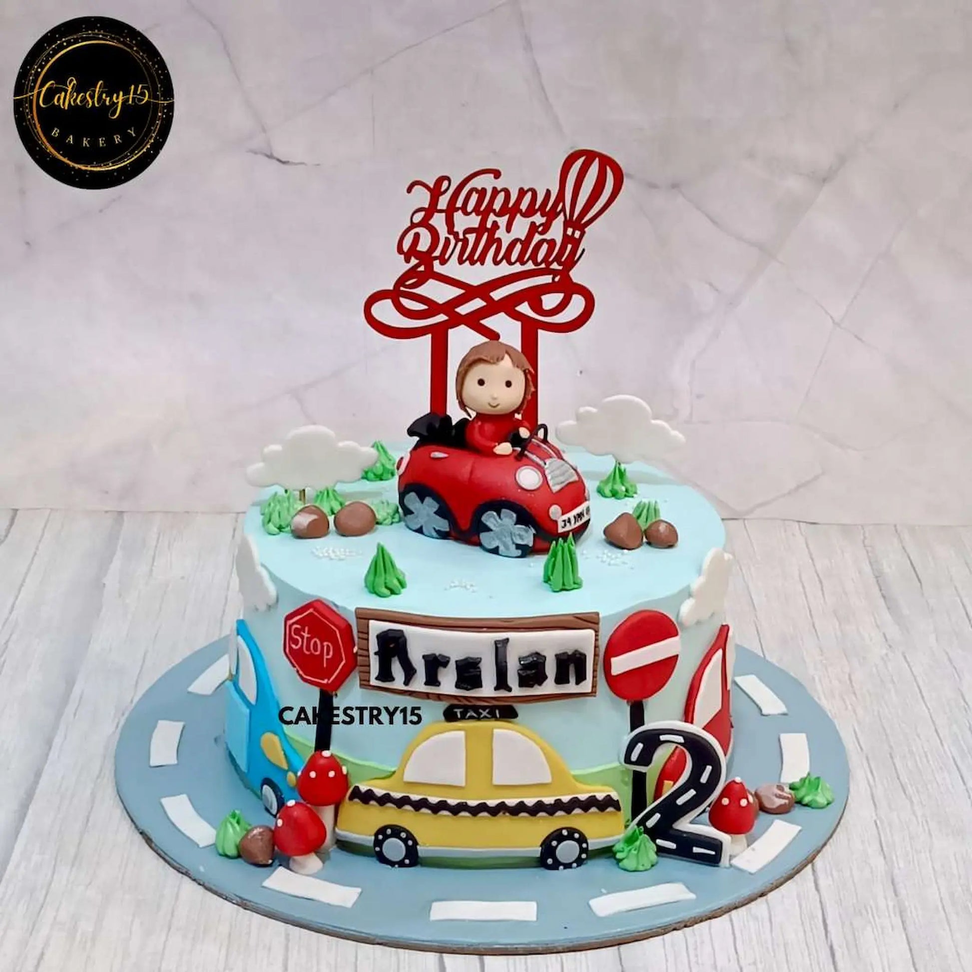 Car-themed birthday cake for Arslan’s 2nd birthday featuring a fondant boy in a 3D car, stop signs, 2D car decorations, and a road base by cakestry15 noida