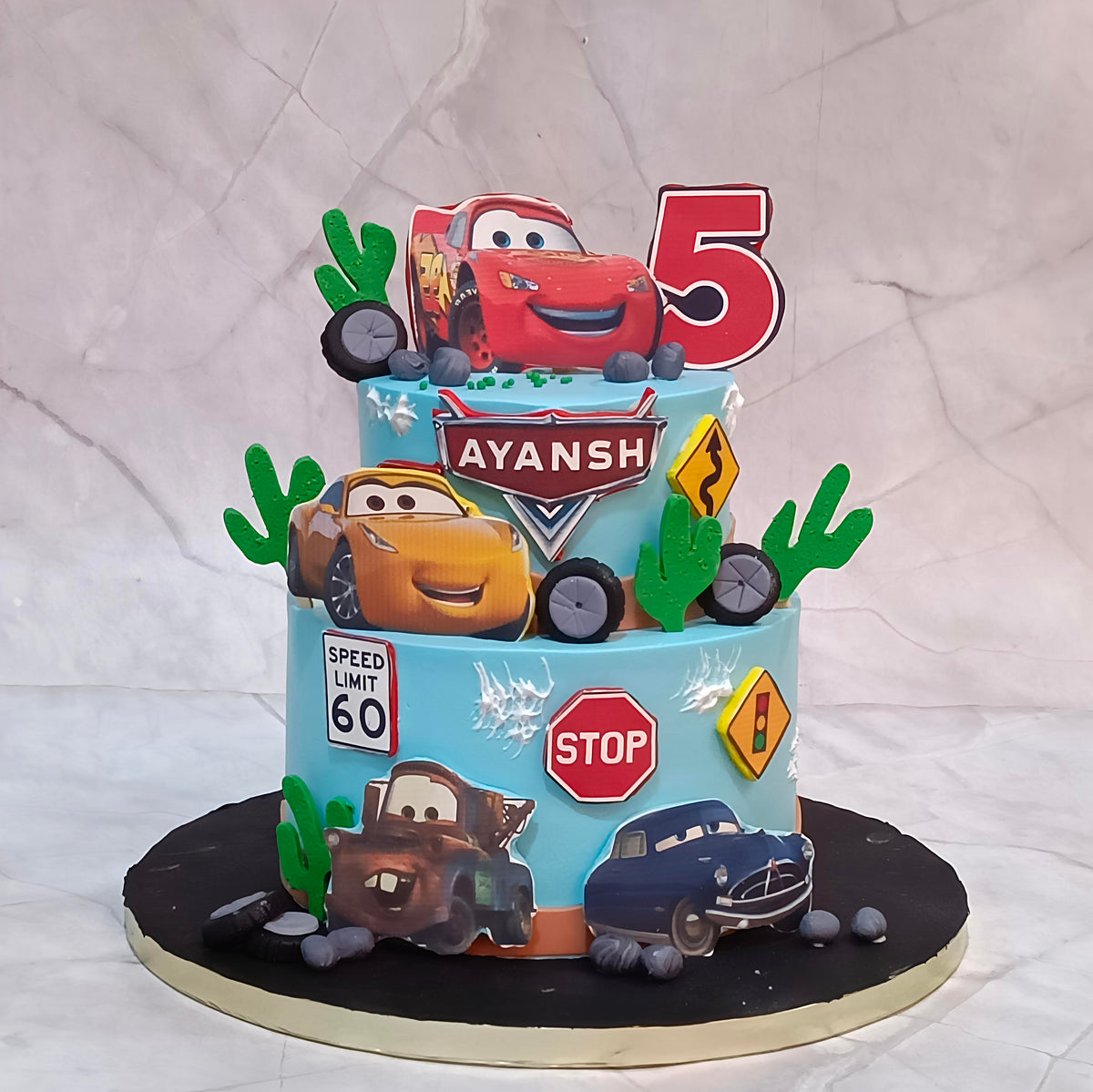 A custom two-tiered Cars-themed birthday cake for Ayansh, decorated with characters Lightning McQueen, Mater, and Rusty, along with road signs, green cacti, and a red number 5 topper, set against a light gray fabric backdrop.