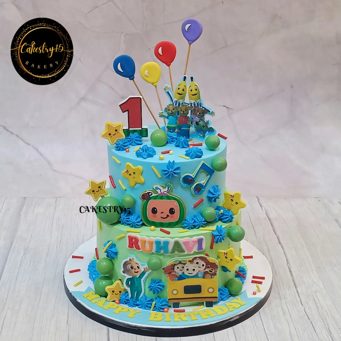 Eggless cocomelon mania first birthday cake with edible characters and balloons for kids birthday celebration in Noida