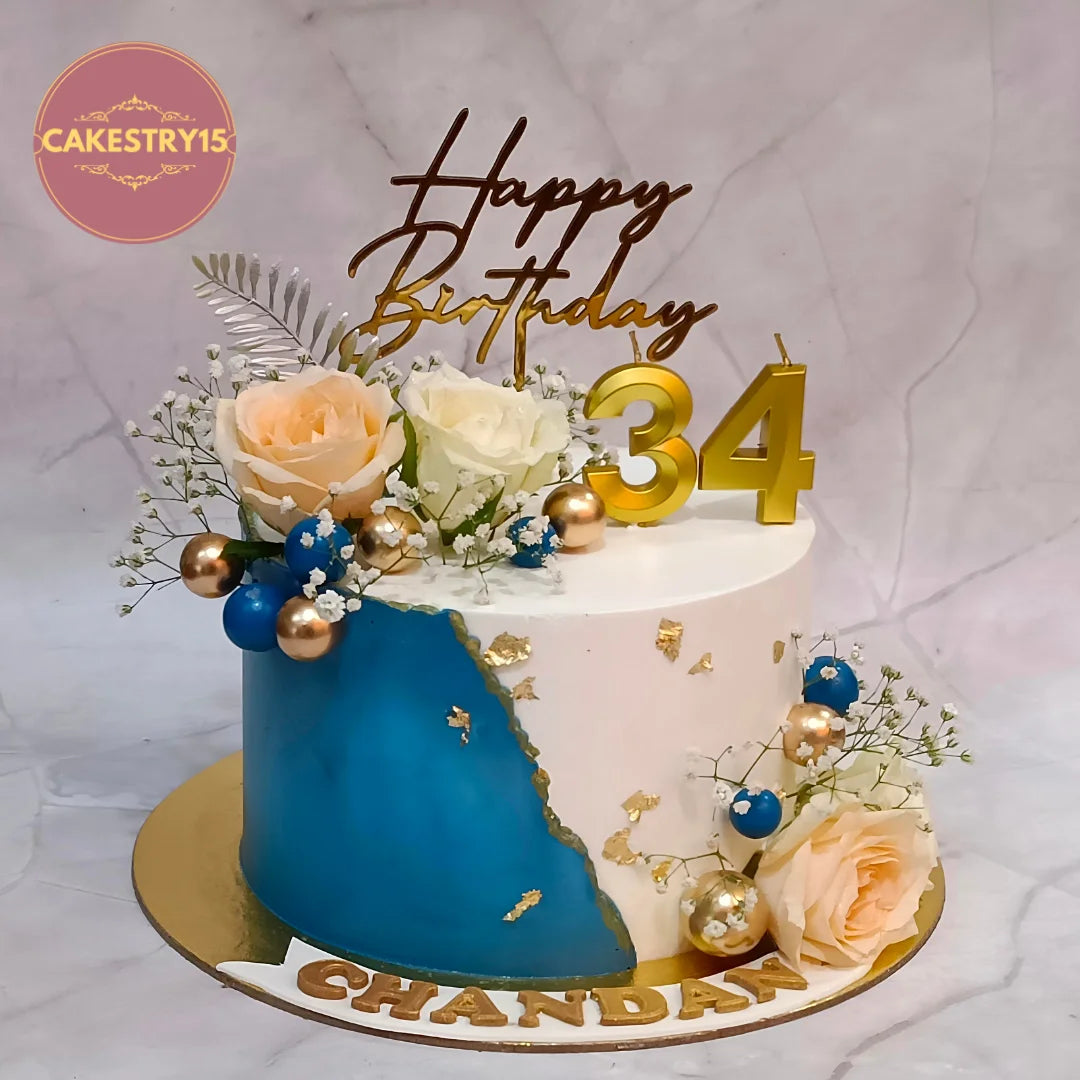 A decorated birthday cake with a blue and white split design, gold leaf accents, peach and white roses, blue and gold ornaments, and a gold 'Happy Birthday 34' topper on a marble background. A Cakestry15 logo is visible in the top left corner.