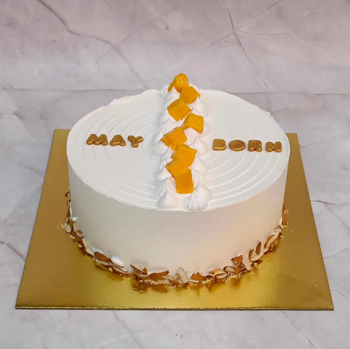 A round birthday cake with white frosting, decorated with a vertical column of orange mango pieces down the center, gold lettering spelling 'MAY BORN' on top, and gold leaf accents around the base, presented on a gold square board against a light marble-patterned background.