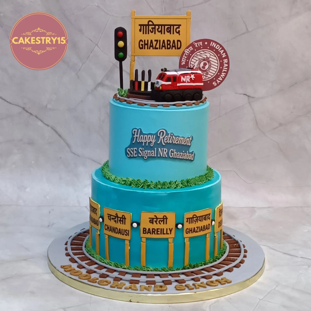 A custom two-tier birthday cake designed for an Indian Railways 'Happy Retirement' SSE Signal NR Ghaziabad. The cake is decorated in light blue with yellow station signposts, brown tracks, green grass, and a red fire engine on top.