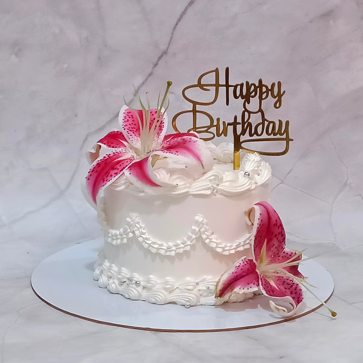White frosted birthday cake with piped borders, a pink and white lily flower and a gold Happy Birthday script topper.