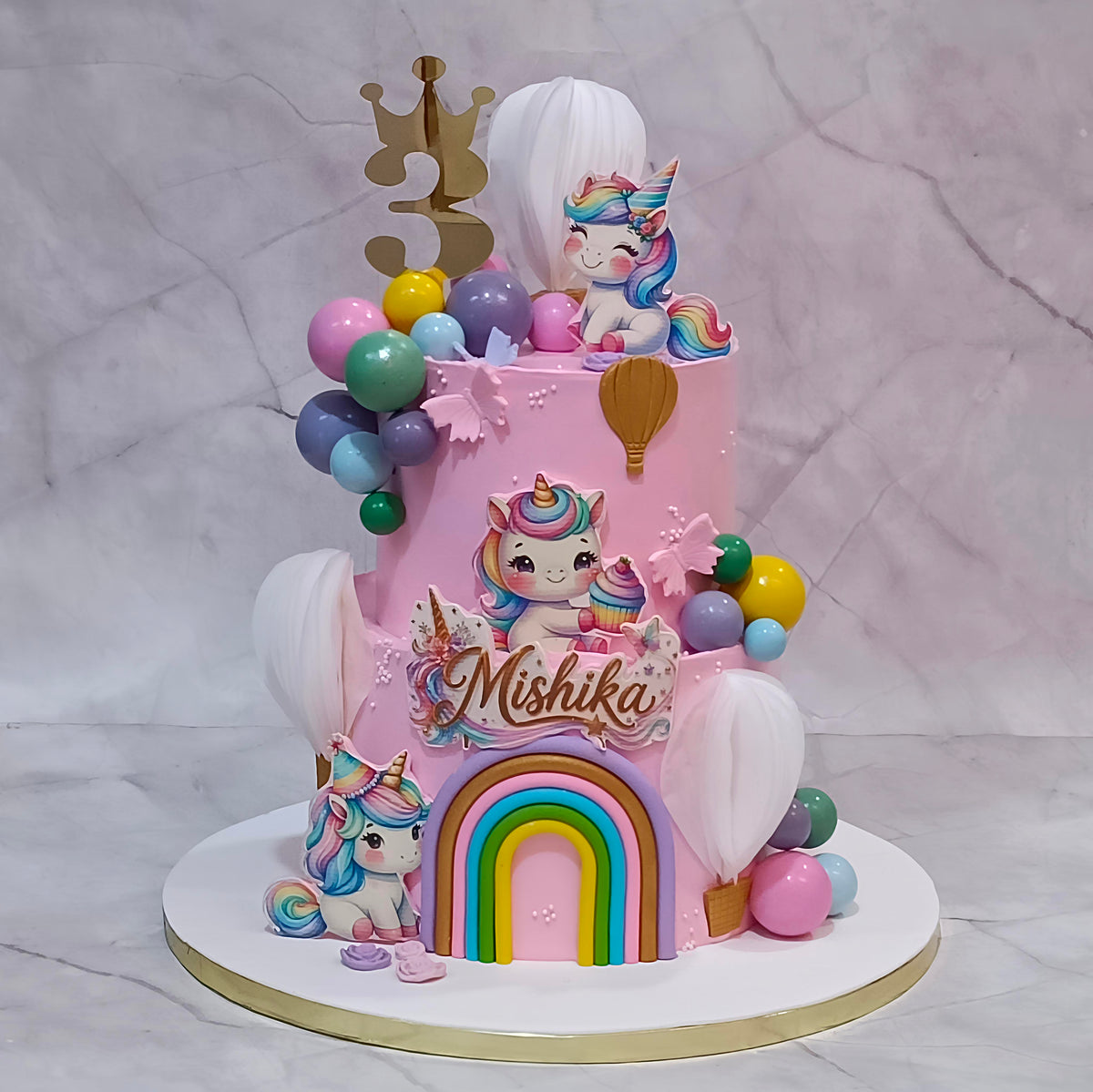 Pink two-tier unicorn birthday cake with edible unicorns, a rainbow arch, pastel balls, air balloon accents and a gold topper.