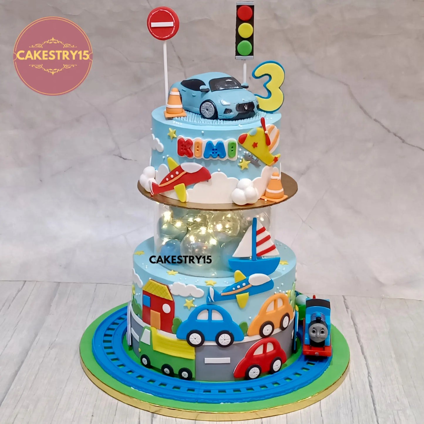 Two-tier birthday cake with spacer,toy cars, traffic lights, and a car on top, featuring the name Kimi and the number 3. Cake is branded with  CAKESTRY15