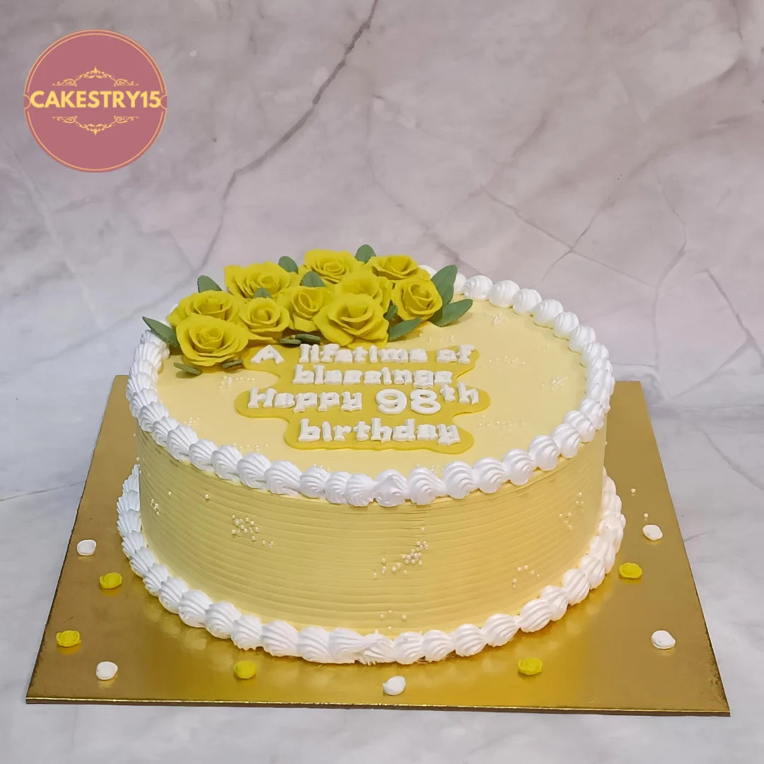A round yellow birthday cake with white piped frosting borders, decorated with yellow rose flowers and green leaves on top. pineapple 2kg cake.