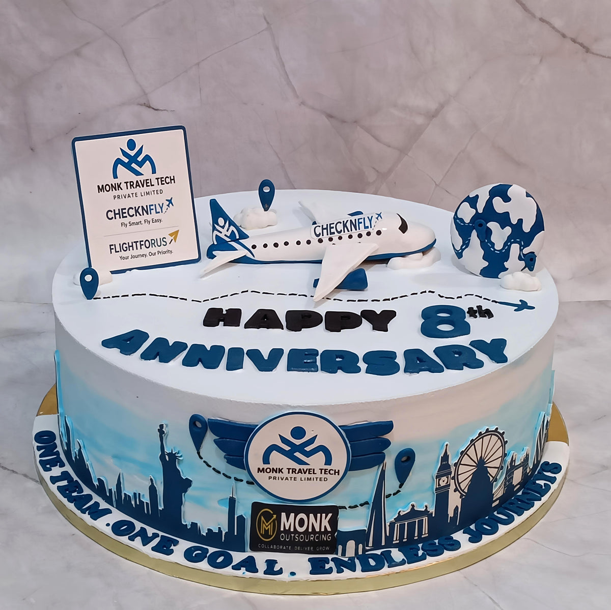 An 8th birthday cake decorated with a white and blue airplane, a world map, a sign reading 'Monk Travel Tech', and 'HAPPY 8th ANNIVERSARY' on the front, set on a marble surface.