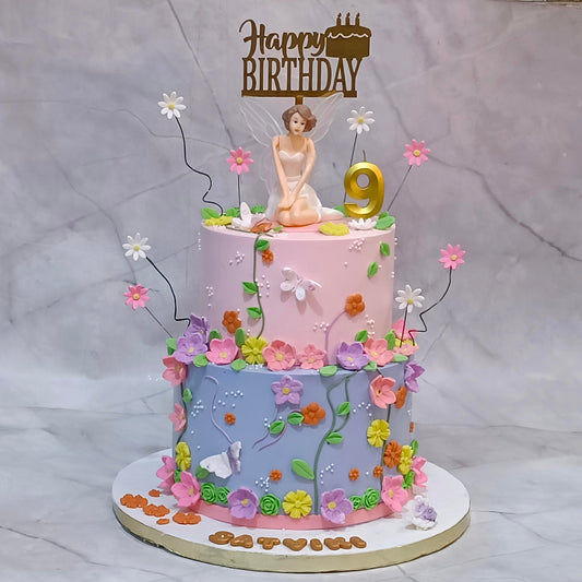 A decorated two-tier birthday cake with a pink top tier and a lavender-blue bottom tier, adorned with colorful flowers, green leaves, butterfly details, a small fairy figurine, and a gold 'Happy Birthday' topper with a gold number 9 candle. 