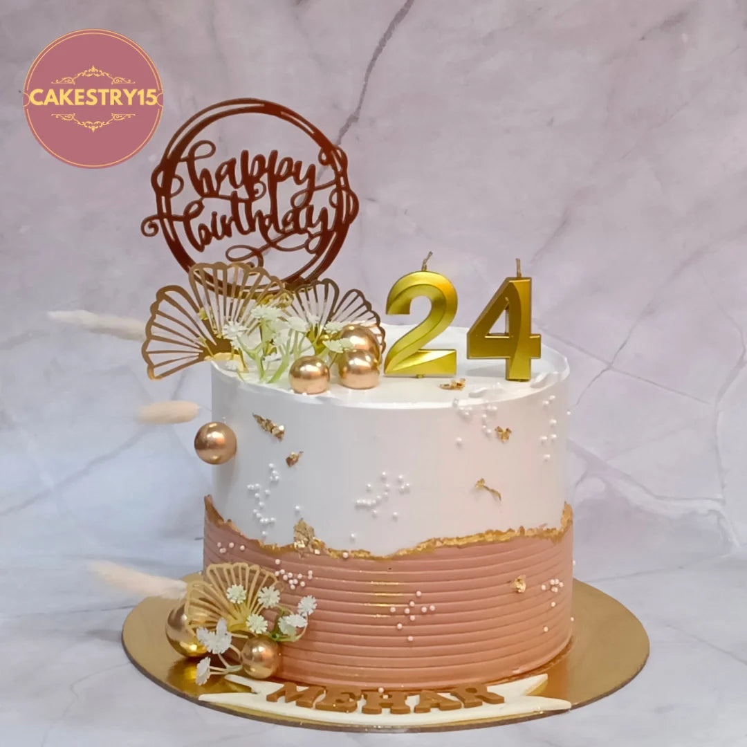 A 24th birthday cake decorated with white and rose gold icing, gold leaf accents, gold 3D numbers, and a brown 'Happy Birthday' script topper, set on a marble background.