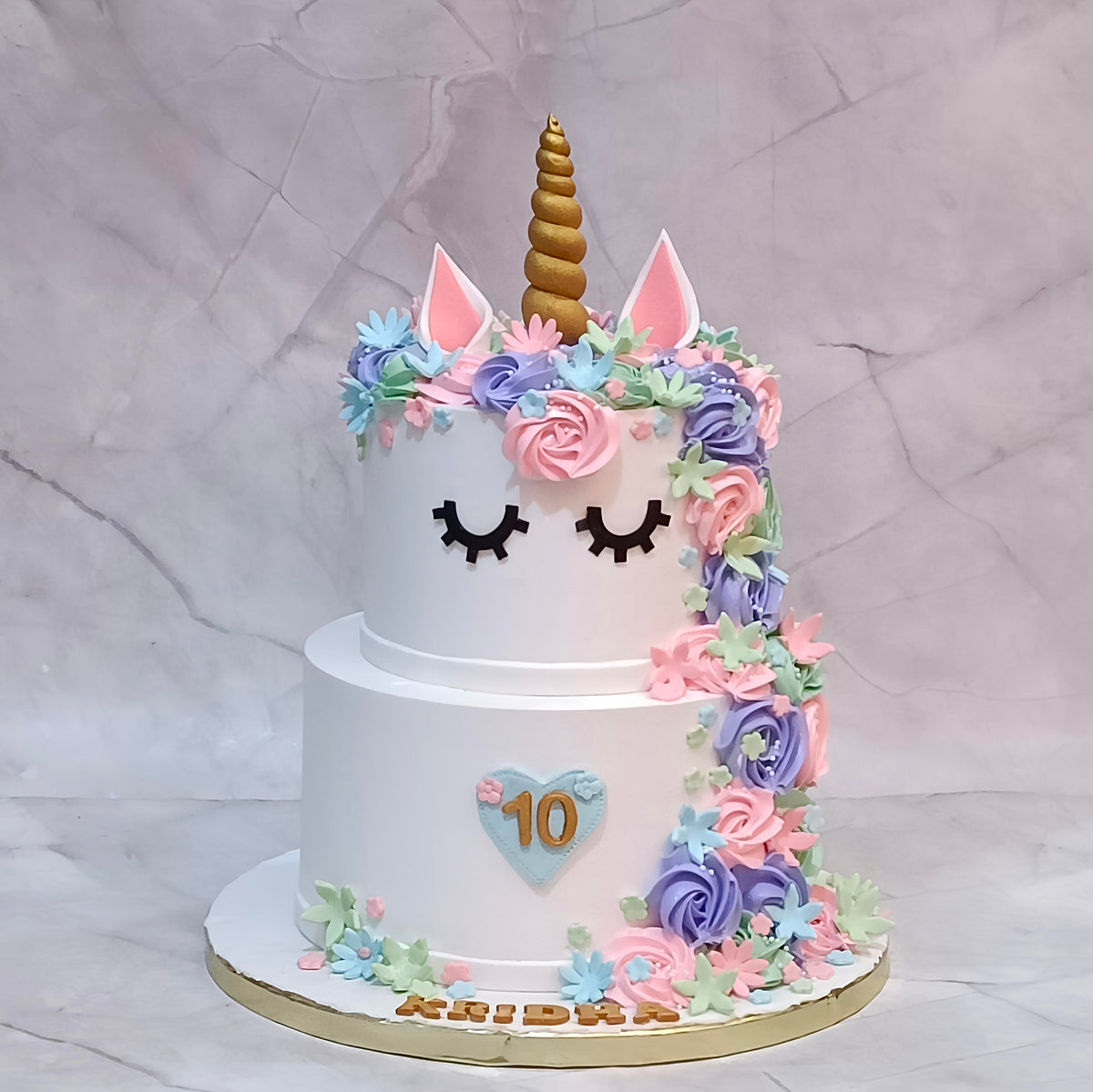 A two-tier white unicorn birthday cake decorated with pink and purple cream flowers, a gold horn, closed black eyes, and a light blue heart with the number 10 on the front.