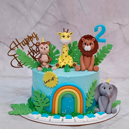 A decorated birthday cake shaped like a jungle safari scene, topped with fondant animal figures including a monkey, giraffe, lion, and elephant, plus a blue number 2 and a 'Happy Birthday' topper on a light gray marble-like background.