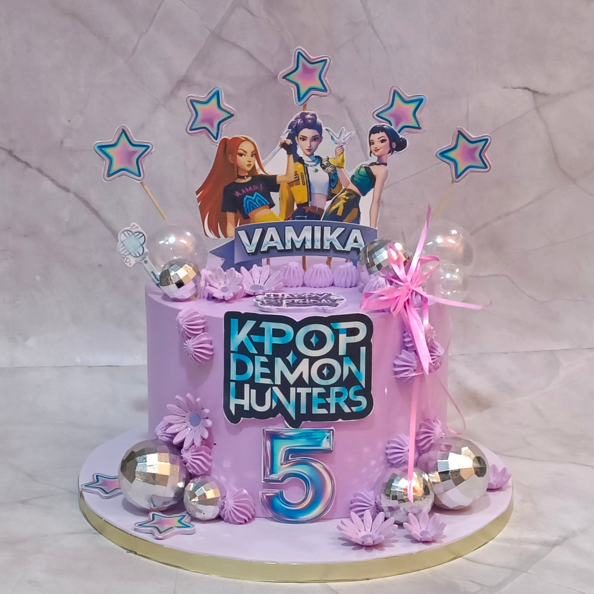A custom purple birthday cake decorated for a K-pop themed celebration. The cake features a large number '5' in blue and pink, a 'K-POP DEMON HUNTERS' logo, character toppers of Lee Min-ha, Sana, and Rosé with the name 'VAMIKA' on a banner, and pink and silver decorations.