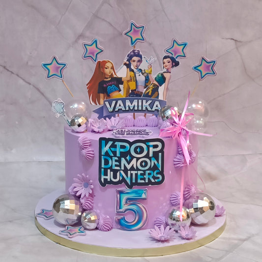A custom purple birthday cake decorated for a K-pop themed celebration. The cake features a large number '5' in blue and pink, a 'K-POP DEMON HUNTERS' logo, character toppers of Lee Min-ha, Sana, and Rosé with the name 'VAMIKA' on a banner, and pink and silver decorations.