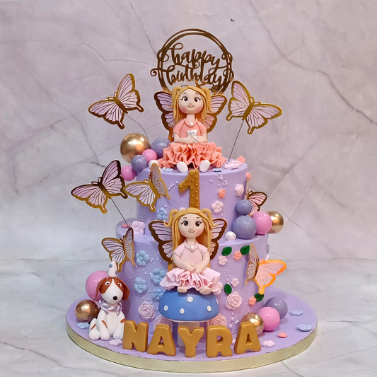 A personalized two-tier birthday cake decorated with fairy figurines, butterflies, pastel flowers, and gold accents. The cake features a purple base, a gold number 1, the name 'NAYRA' in gold letters, and a 'Happy Birthday' topper