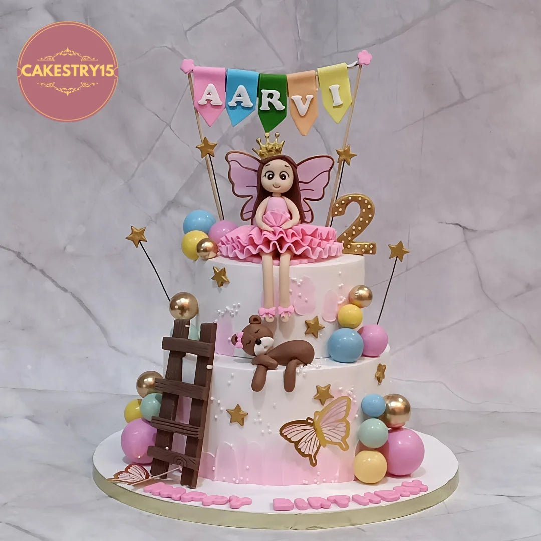 A custom two-tier birthday cake decorated for a girl named Aarvi. The cake features pink and white tiers, gold stars, pastel balloons, a brown teddy bear, a butterfly, a small fairy figurine, a chocolate ladder, and a colorful 'AARVI' banner on top.
