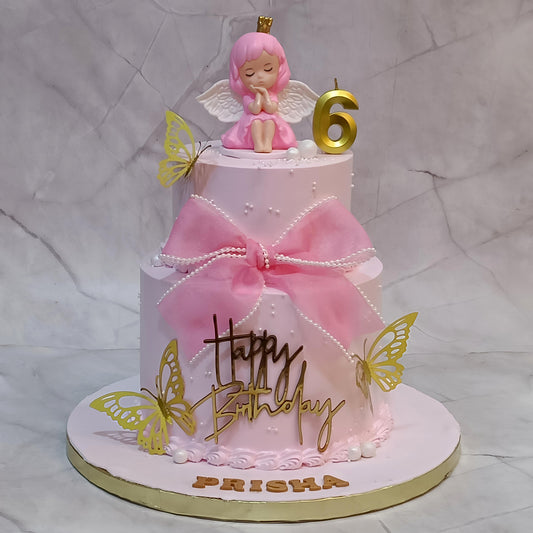 A pink fairy themed two-tier birthday cake decorated with a large pink bow, gold script reading 'Happy Birthday,' white pearl-like accents, gold butterflies, and a pink angel figurine with a gold number 6 candle on top. The cake is displayed on a round cake board with the name 'PRISHA' at the base, set against a light marble-patterned backdrop.