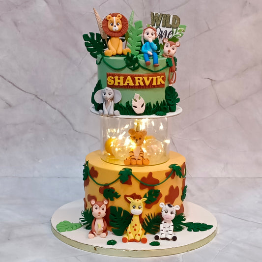 A custom three-tiered safari-themed birthday cake decorated with animal figurines, green leaves, and a clear light-up center. The top tier is green with a lion, monkey, and 'WILD ONE' topper, and the middle tier is yellow with a giraffe and elephant figurine.
