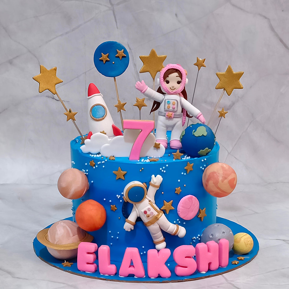 Astronauts and planets space theme cake with blue icing, two fondant astronaut figurines, a rocket and golden stars.