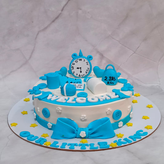 A custom baby shower cake decorated in blue and white with fondant details, featuring a clock, diaper, stethoscope, and a large bow on top, displayed on a white marble-patterned background.