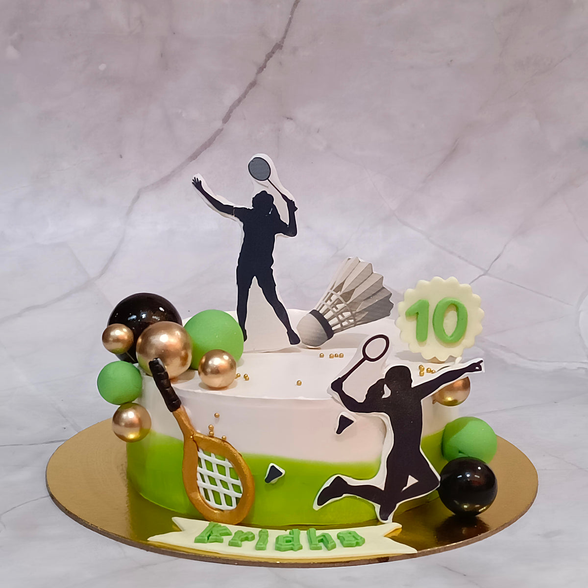 A custom 10th birthday cake decorated with badminton-themed elements, including silhouettes of players, a shuttlecock, a racket, green and gold balloons, and a '10' topper, on a marble-patterned background.
