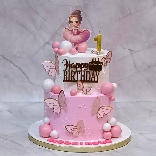 Two-tier ballerina theme first birthday cake with an edible ballerina image, number 1 candle, faux butterflies and decorative balls.