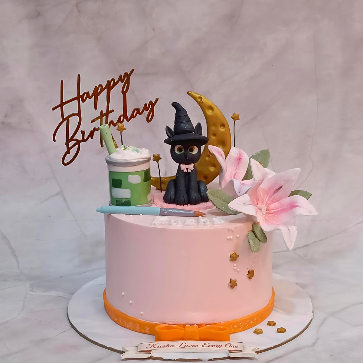 A pink frosted birthday cake with a black cat figurine wearing a witch hat, a gold crescent moon, pink lily flowers, a green cup decoration, and a brown 'Happy Birthday' script topper on a white board.