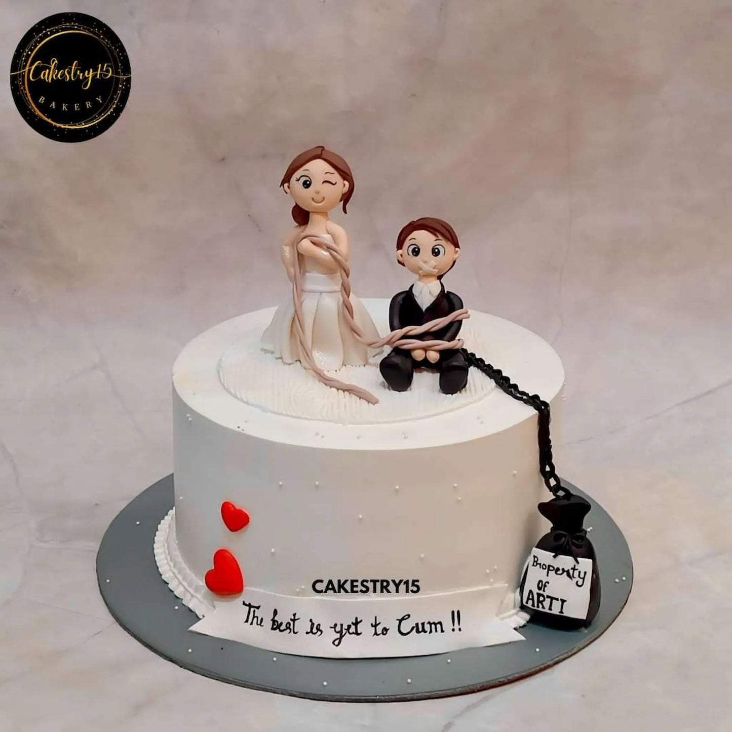 A fun and bold 2kg Black Forest bachelorette cake featuring a bride holding a groom tied up with chains and a bag that says “Property of Arti by cakestry15,best bakery noida