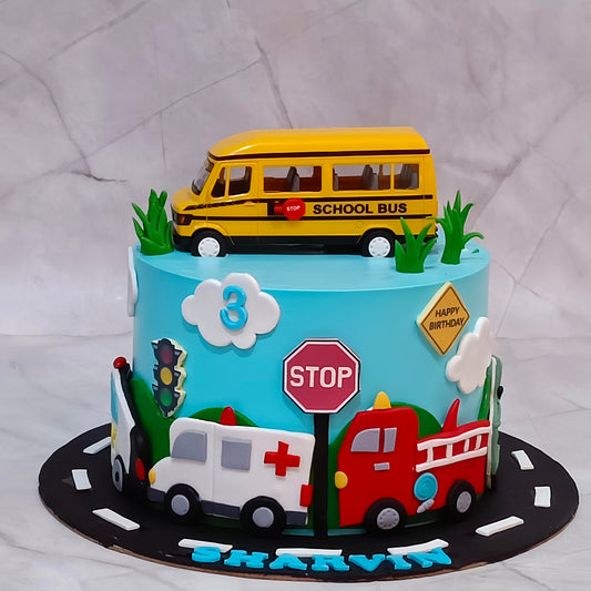 A custom bus theme cake for a 3-year-old birthday  decorated with school bus, ambulance, fire truck, stop sign, traffic light, clouds, and green grass, topped with a yellow school bus topper. The cake is personalized with the name 'SHARVIN' in blue icing on a black road-patterned base.