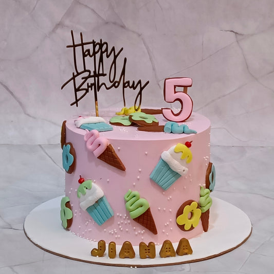 A pink frosted birthday cake with ice cream and cupcake decorations, topped with a 'Happy Birthday' script topper and a large number 5 topper. The cake is decorated with colorful fondant ice cream cones, cupcakes, flowers, and the name 'JIANAA' spelled out in letters on the front board.