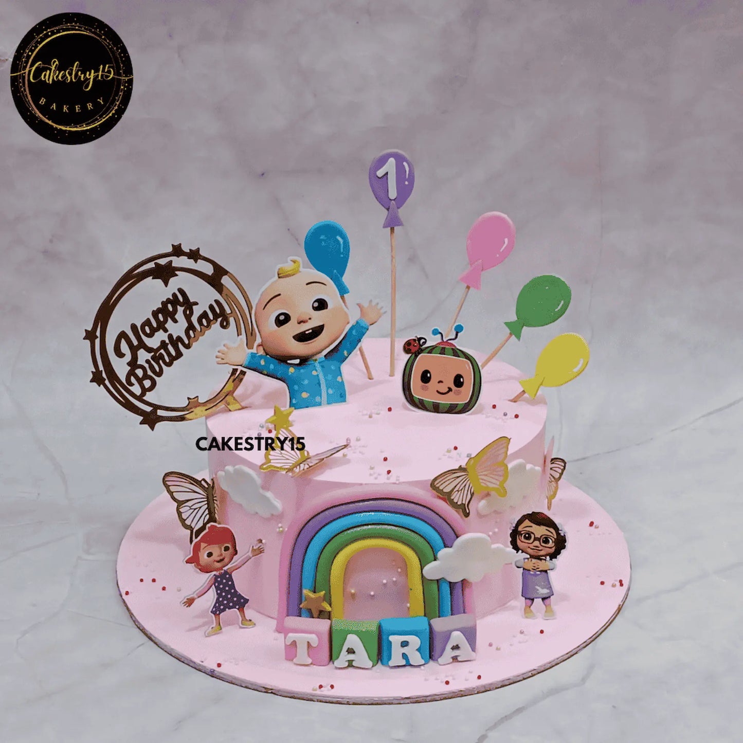 Cocomelon rainbow first birthday cake for kids in Noida with pink base, fondant rainbow and character toppers, eggless custom cake