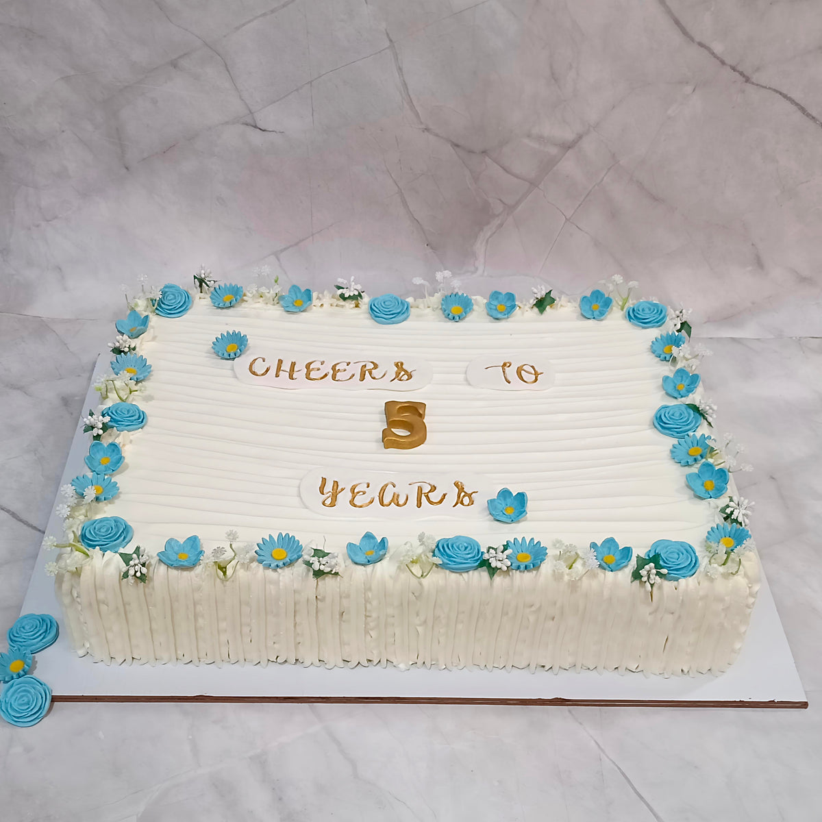 A rectangular  corporate anniversary cake decorated with white frosting, blue and white flower accents, and gold lettering that reads 'Cheers to 5 years'.