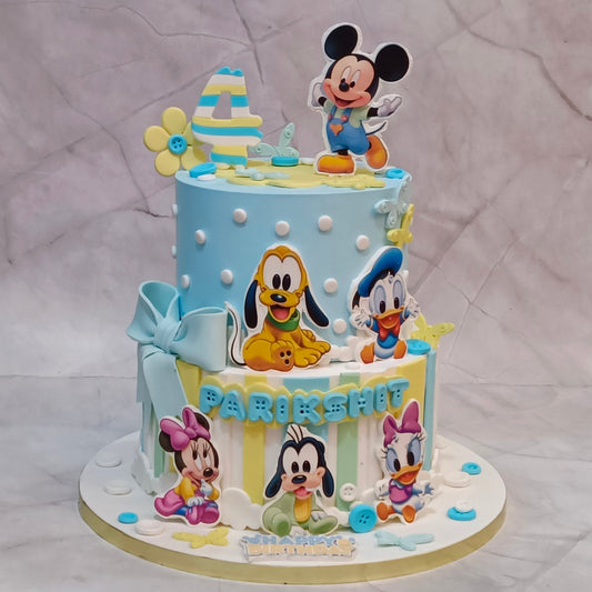 A decorated two-tier birthday cake featuring Disney characters including Mickey Mouse, Pluto, Donald Duck, Minnie Mouse, and Daisy Duck, with a light blue and yellow color scheme and the name on the front.