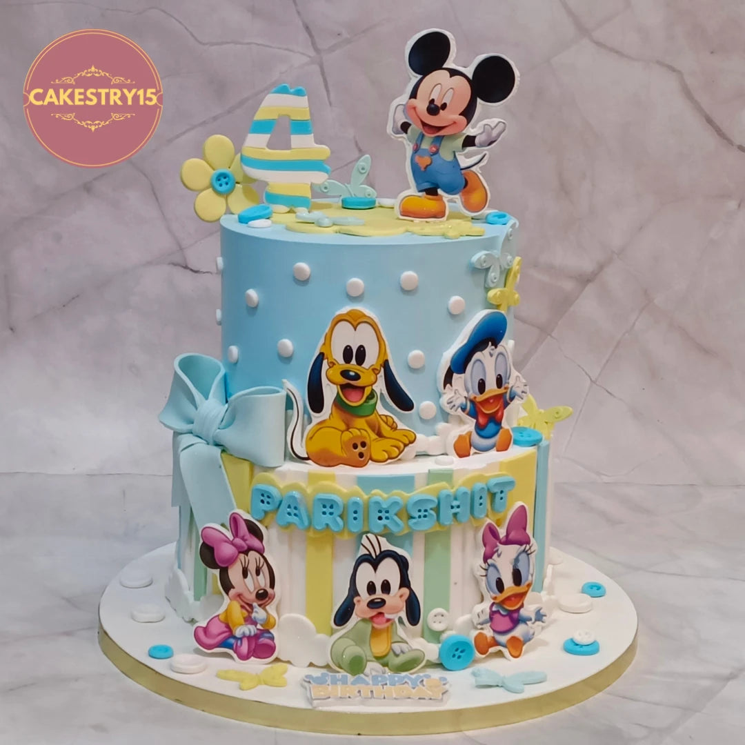 A decorated two-tier birthday cake with Disney characters, featuring Mickey Mouse on top, Pluto and Donald Duck on the upper tier, and Minnie, Daisy, and Pluto on the lower tier. The cake is light blue and yellow with white polka dots and a large light blue bow.