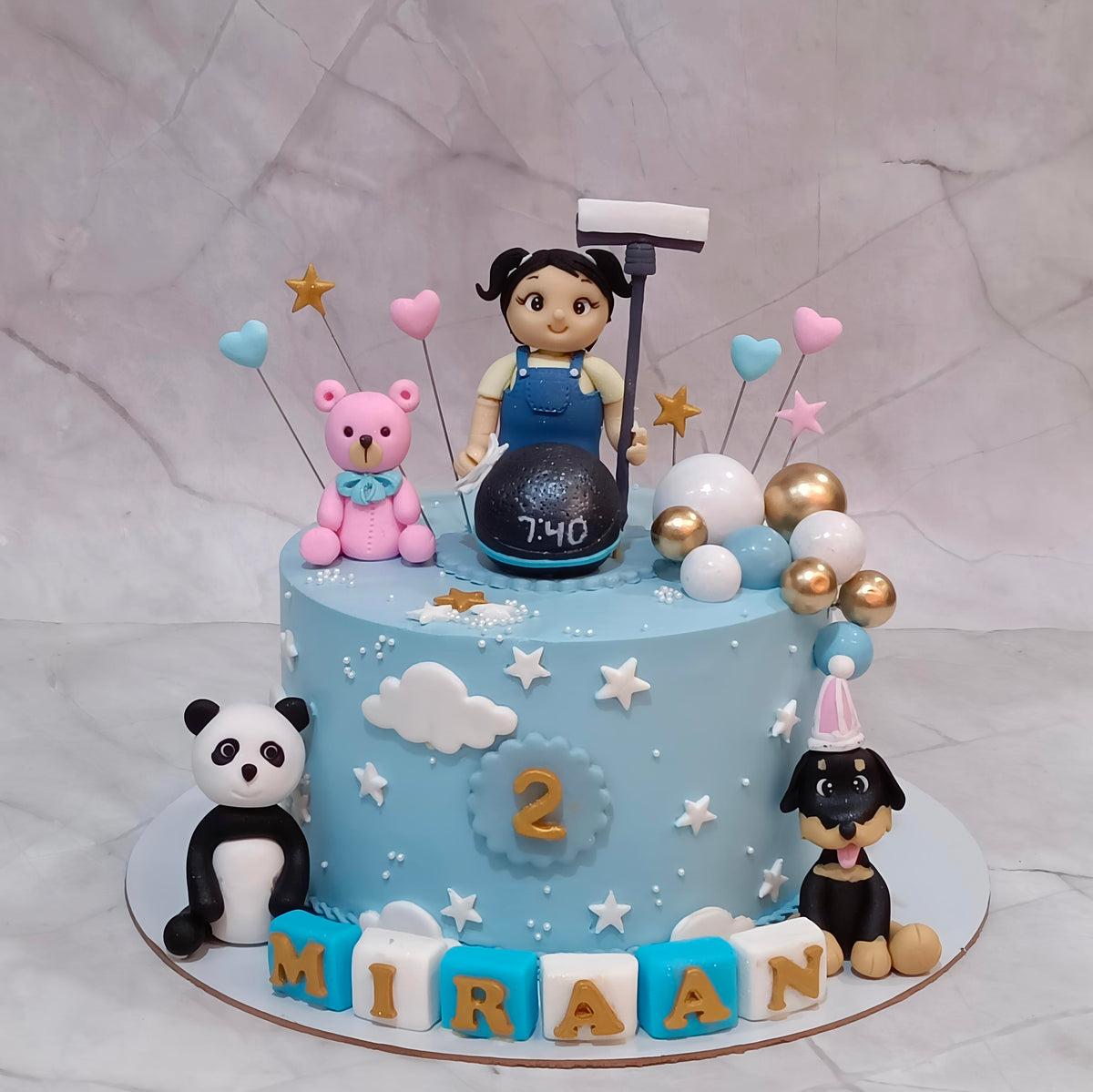 A decorated light blue birthday cake with a child's room theme, featuring a cartoon girl topper, a pink teddy bear, a panda figurine, a dog figurine, balloons, and letter blocks spelling 'MIRAAN' on a marble background.