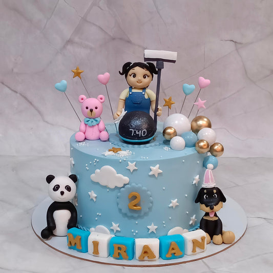 A decorated light blue birthday cake with a child's room theme, featuring a cartoon girl topper, a pink teddy bear, a panda figurine, a dog figurine, balloons, and letter blocks spelling 'MIRAAN' on a marble background.