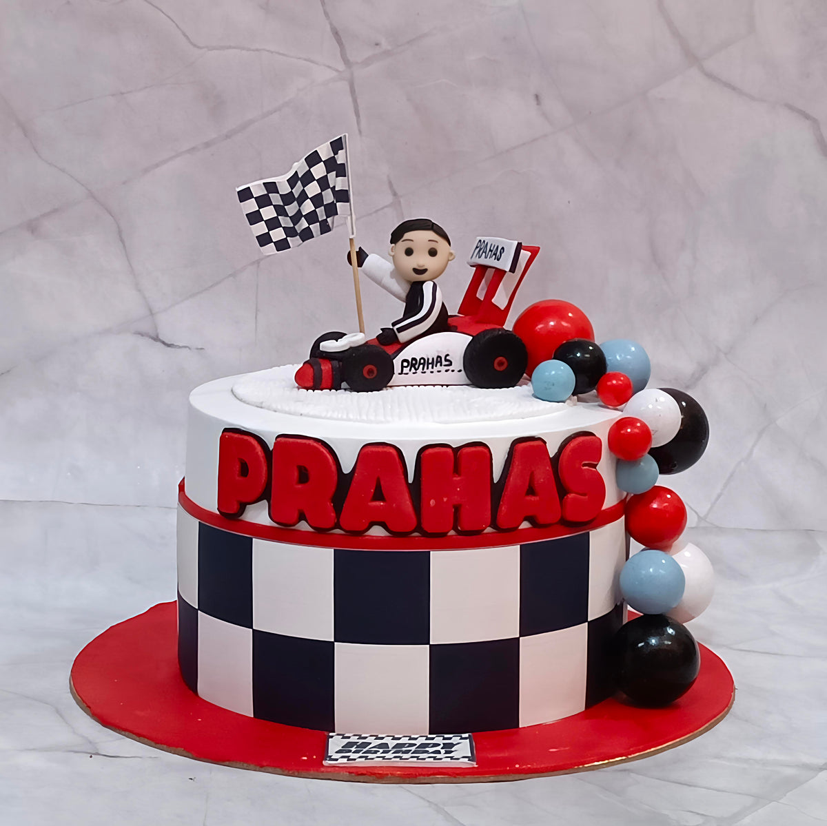 F1 race car theme birthday cake with a kid figurine, chequered base, racing car and black, white and red faux balls.