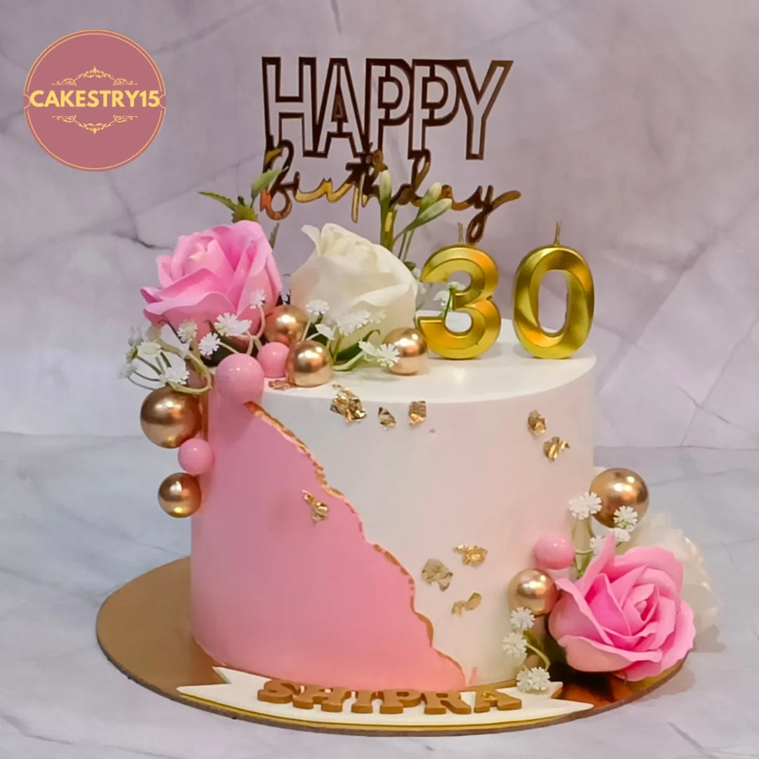 A decorated birthday cake with pink and white frosting, gold leaf accents, pink and white roses, gold baubles, and gold number 30 toppers on a gold cake board. 