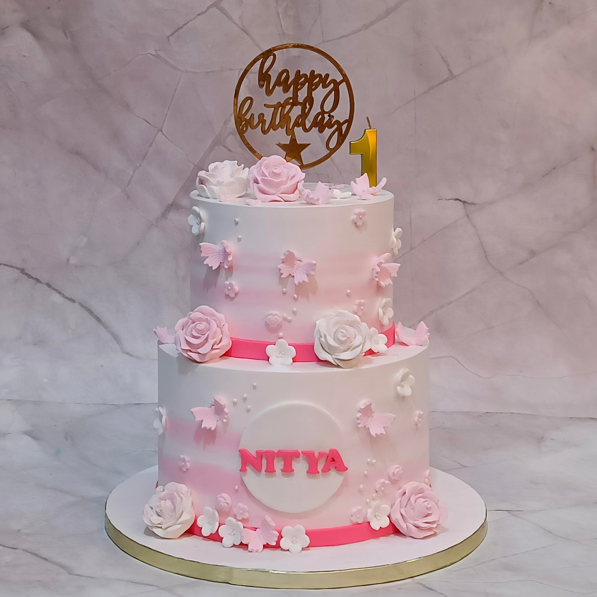 A two-tiered custom floral first  birthday cake with a pink and white ombre gradient, decorated with 3D roses, butterflies, and flowers. The cake features a gold 'happy birthday' topper with a gold '1' candle, and the name 'NITYA' written in pink on the front.
