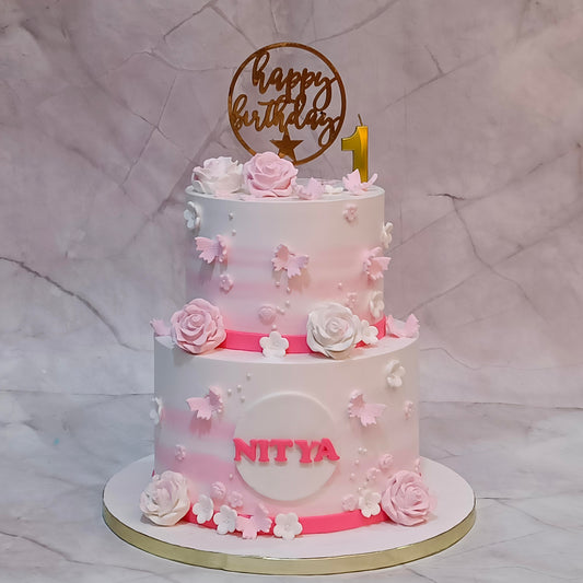 A two-tiered custom floral first  birthday cake with a pink and white ombre gradient, decorated with 3D roses, butterflies, and flowers. The cake features a gold 'happy birthday' topper with a gold '1' candle, and the name 'NITYA' written in pink on the front.