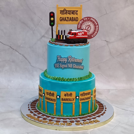 A custom two-tier birthday cake designed for an Indian Railways 'Happy Retirement' SSE Signal NR Ghaziabad. The cake is decorated in light blue with yellow station signposts, brown tracks, green grass, and a red fire engine on top.