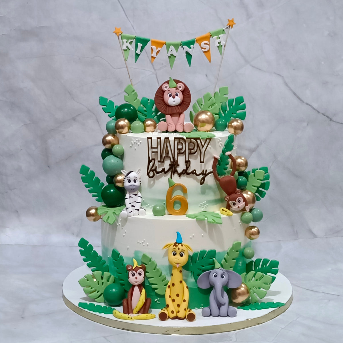 A decorated two-tier birthday cake with jungle-themed fondant decorations, including green leaves, gold and green beads, animal figurines, and a lion topper. The cake is topped with a banner  and a gold number 6, with 'HAPPY Birthday' written on the front.