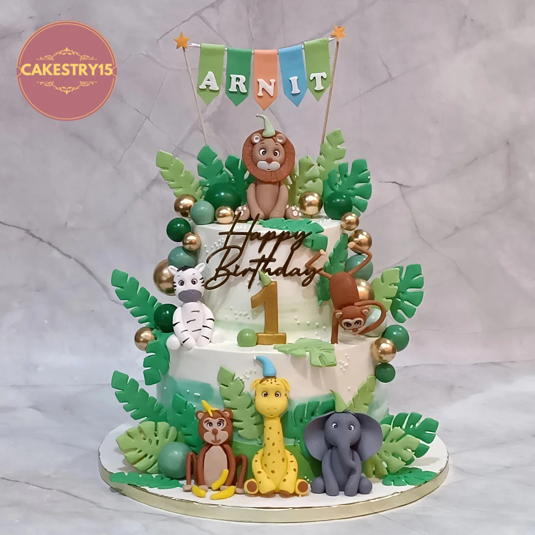 A custom two-tiered birthday cake decorated with safari animal figures, green leaves, gold accents, and a 'Happy Birthday' topper, displayed on a marble surface.