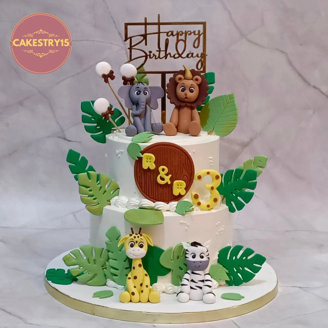 A two-tiered white birthday cake decorated with safari-themed fondant figures, including a lion, elephant, giraffe, and zebra, surrounded by green monstera leaves. The cake features a wooden 'Happy Birthday' sign on top and a round brown plaque with the names 'R & R' and the number '3' in yellow.