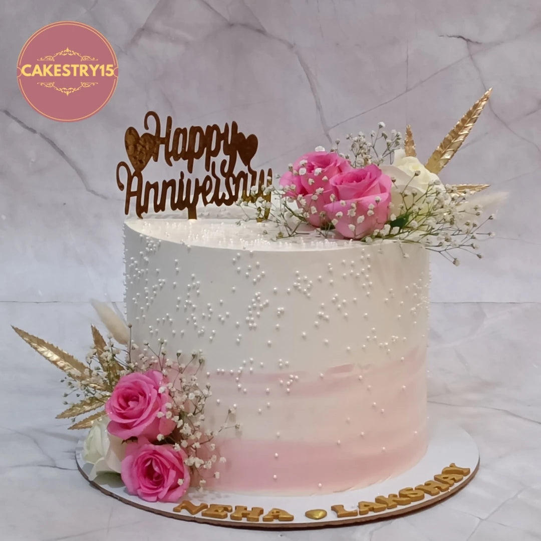 Eggless first anniversary cake featuring fresh roses, baby breath, white pearls, personalised names, and an anniversary topper, available in Noida.
