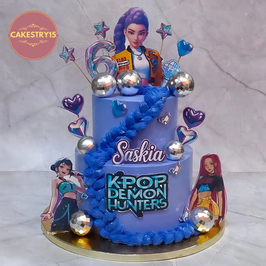 A custom two-tier birthday cake themed around the anime K-pop group 'Demon Hunters' and featuring the character Saskia. The cake is decorated with light blue and royal blue frosting, silver disco ball toppers, heart-shaped and star-shaped cake toppers, and character cutouts.