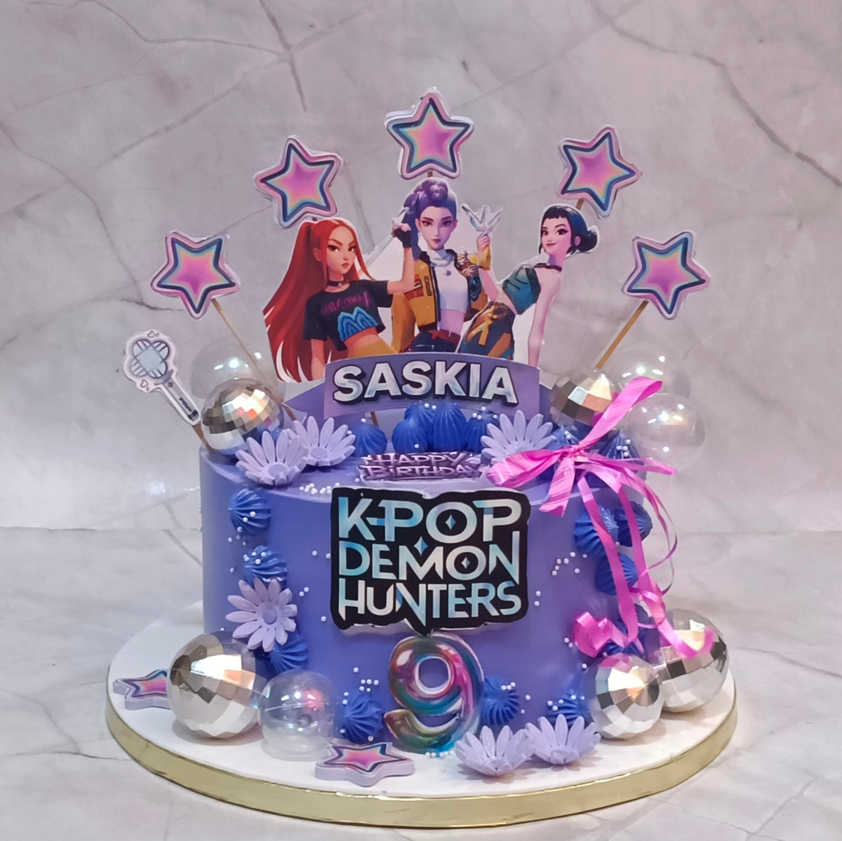 A custom purple birthday cake for a K-pop fan named Saskia, decorated with 'K-POP DEMON HUNTERS' text, character cutouts, star toppers, and silver disco ball accents, set on a marble-patterned background.